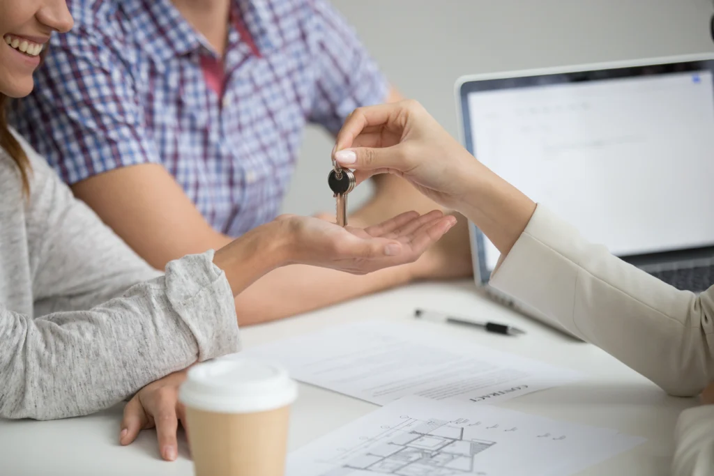a couple receives a key for their new home as they sit at a desk with house plans covering it.